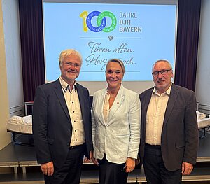 Eine Frau zwischen zwei Männern vor dem Schriftzug "100 Jahre DJH Bayern".