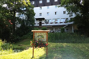 Haus im Hintergrund, davor Grün und ein Teich mit Hinweistafel