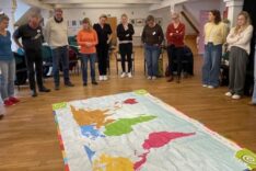 Eine Gruppe Erwachsener steht im Kreis um eine Weltkarte, die auf dem Boden ausgebreitet liegt. Die Referentin erklärt das Weltspiel, dass in der nächsten halben Stunde gespielt wird.