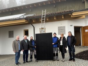 Menschen stehen vor einem Haus, in der Mitte halten sie ein Solarmodul.