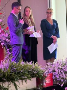 Ein Mann und zwei Frauen neben Blumengestecken, er spricht in ein Mikrofon