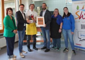 Drei Frauen und drei Männer mit "Bio"-Zertifikat in einem Bilderrahmen und einem Geschenkkorb.