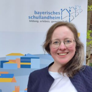 Eine Frau mit Brille vor dem Logo des Bayerischen Schullandheimwerks.